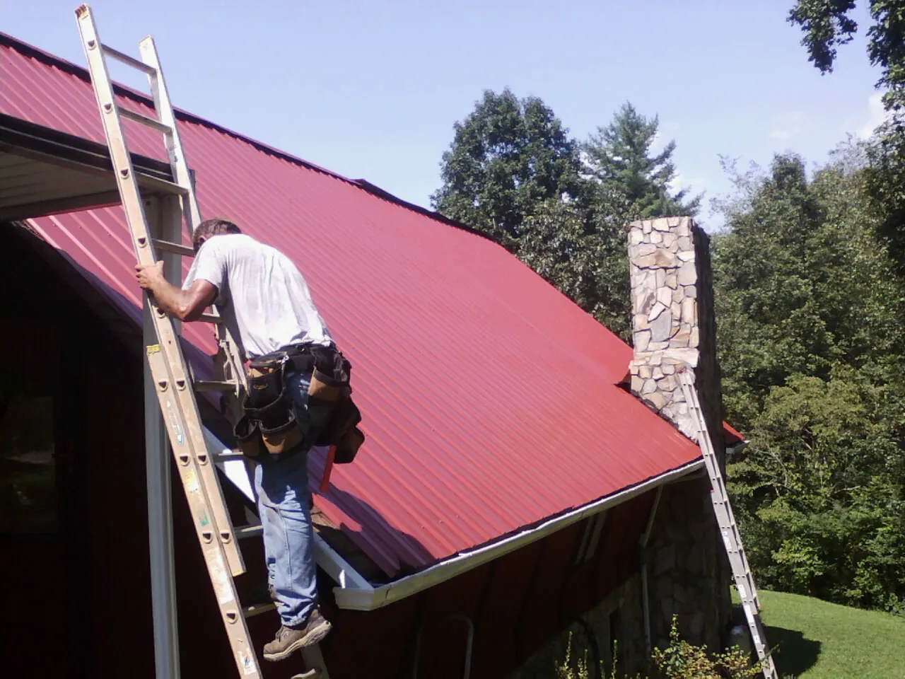 Roofer on a ladder beside a red metal roof and stone chimney — Soffit & Fascia Repair by Roofing Brothers in Sioux City